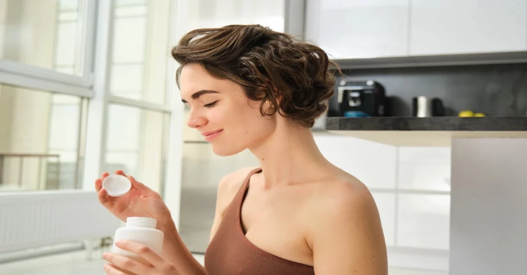 Une femme en débardeur marron tient un flacon blanc, présentant un complément alimentaire pour femmes dans sa cuisine moderne