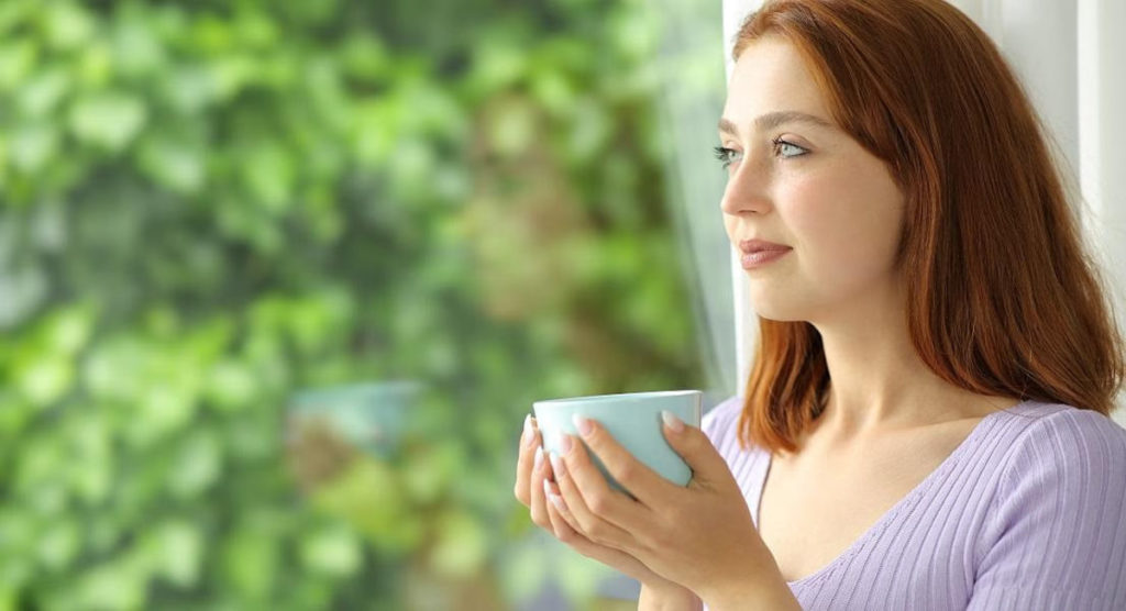 Woman holds a blue mug and gazes out a window, reflecting the Myo-Inositol benefits she’s experienced in her wellness journey